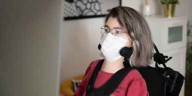 A power wheelchair user wearing a mask sits in front of a computer monitor.
