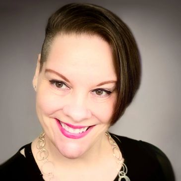 A woman with brown hair in an asymetrical haircut, pink lipstick and geometric neclace smiles.