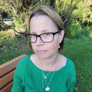 A woman with blond and brown hair wears a green top and glasses. She has two necklaces on.