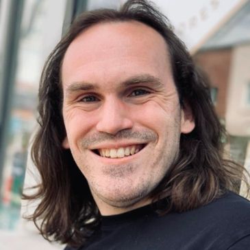 A smiling man with shoulder-length brown hair and a black shirt smiles into the camera.