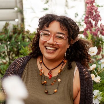 A smiling woman with brown skin and hair is wearing glasses, a green top and several necklaces.