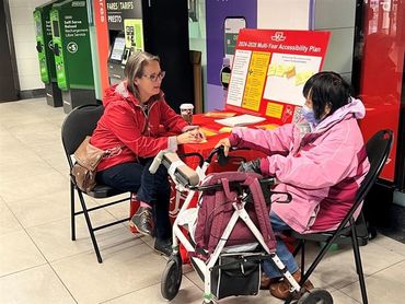 A woman in a red jacket talks to a woman in a pink jacket who has a walker.