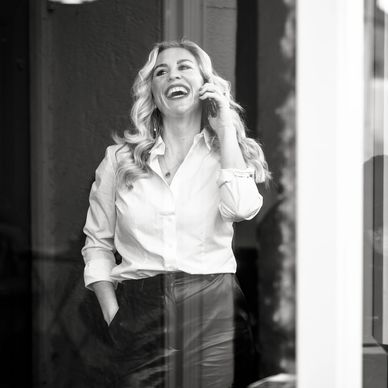 Woman joyfully talking on phone, dressed in white shirt and dark pants.