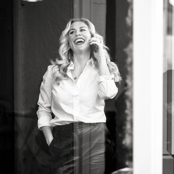 Woman joyfully talking on phone, dressed in white shirt and dark pants.