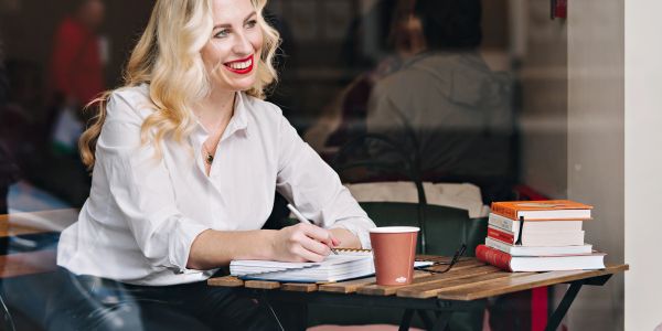A woman with blonde hair writing in a notebook at a café table.