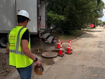 Worker in safety gear near a manhole and traffic cones.