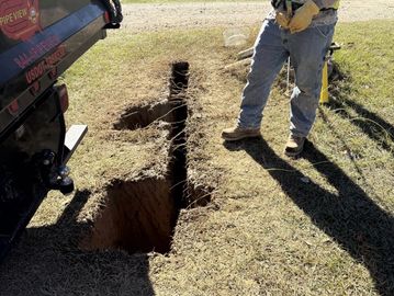 A person standing near a freshly dug trench in grassy ground.