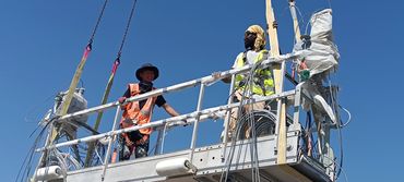 Two construction workers stand on a suspended platform against a clear blue sky.