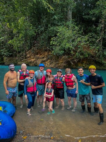 A group of people in life jackets and helmets standing in shallow water by a forested riverbank.