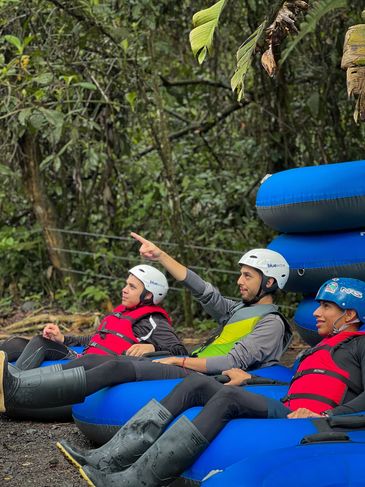Three young men in helmets and life jackets sitting on blue inflatable rafts in a forest.