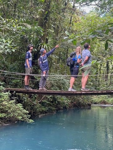 Four people stand on a wooden bridge over blue water in a lush forest.