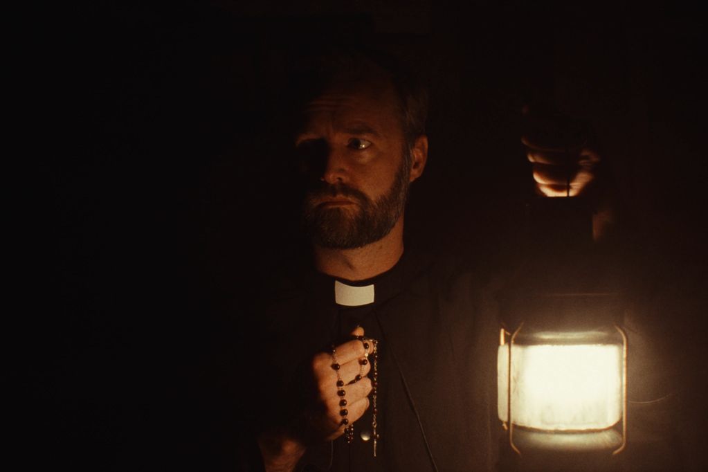 A priest holding a lantern and rosary in darkness.