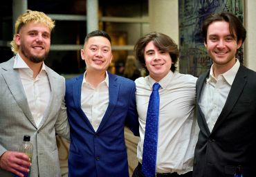 Four men in suits smiling at the camera at a social event.