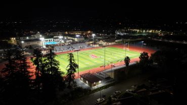 Night view of a lit sports field with surrounding buildings and trees.