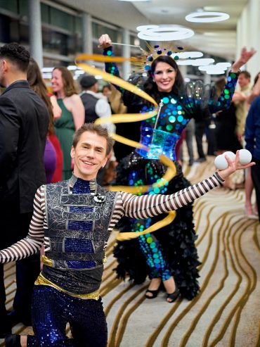 Two performers in colorful costumes entertain a lively crowd indoors.