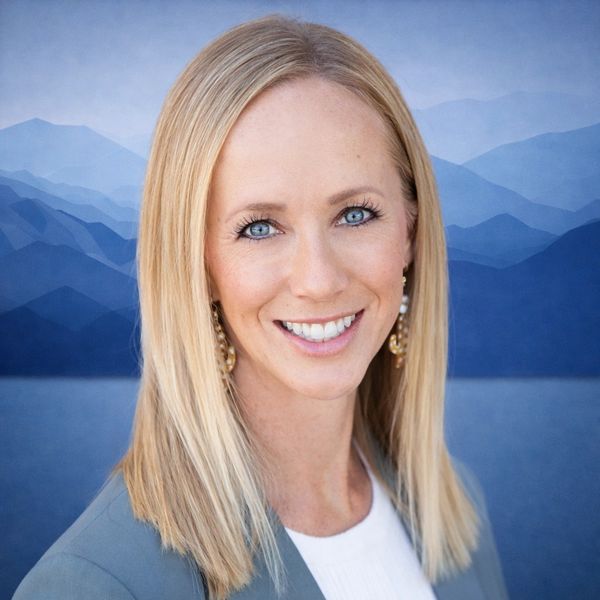 Smiling blonde woman with blue eyes against a blue mountain background.