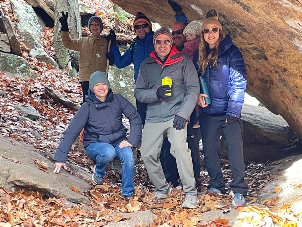 A group of friends posing under a large rock in a forest during autumn.