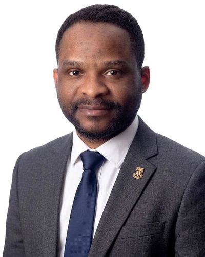 Professional portrait of Dr Godwin Ukeje in a suit and tie against a grey background.