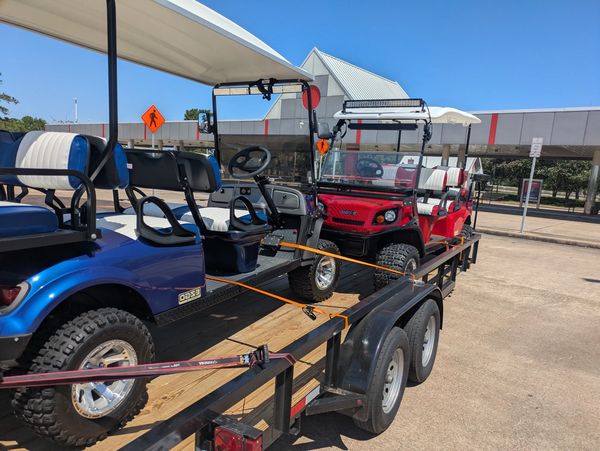 Two golf carts, one blue and one red, secured on a black trailer in a parking lot.