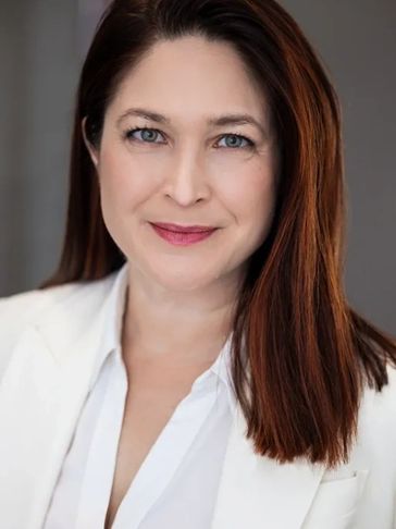 Professional woman with red hair and a white blazer smiling gently.