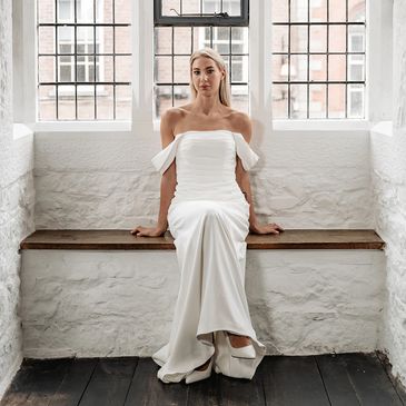 A woman in an elegant white dress sitting on a wooden bench by a window.