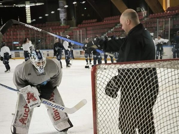 Hockey coach training goalie on ice rink with players in background.