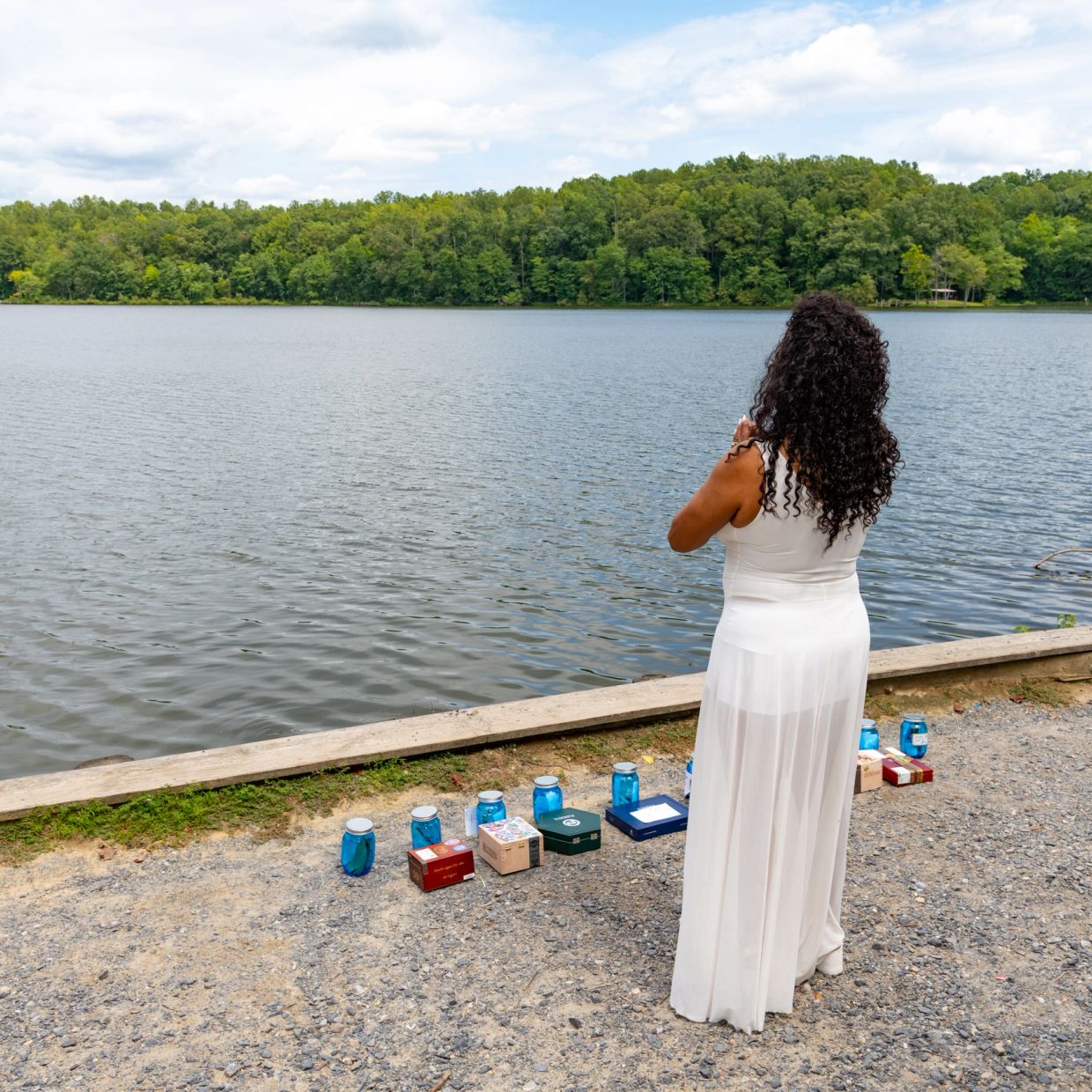 Woman in white dress standing by a lakeside with blue jars and boxes.