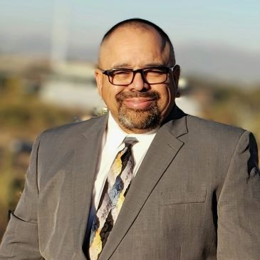 Man in a suit and glasses smiling outdoors.