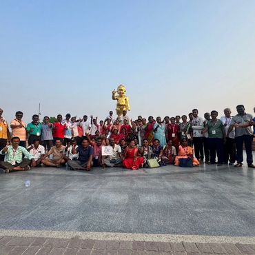 Large group posing with thumbs up in front of a golden statue outdoors.
