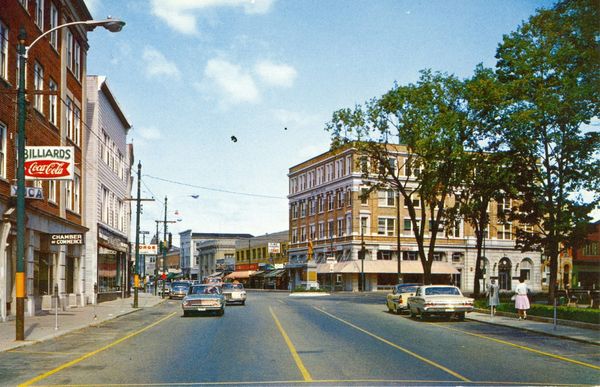 A vintage downtown street scene with classic cars and storefronts under a blue sky.