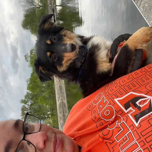 Person in orange hoodie holding a small black and tan dog near a body of water.