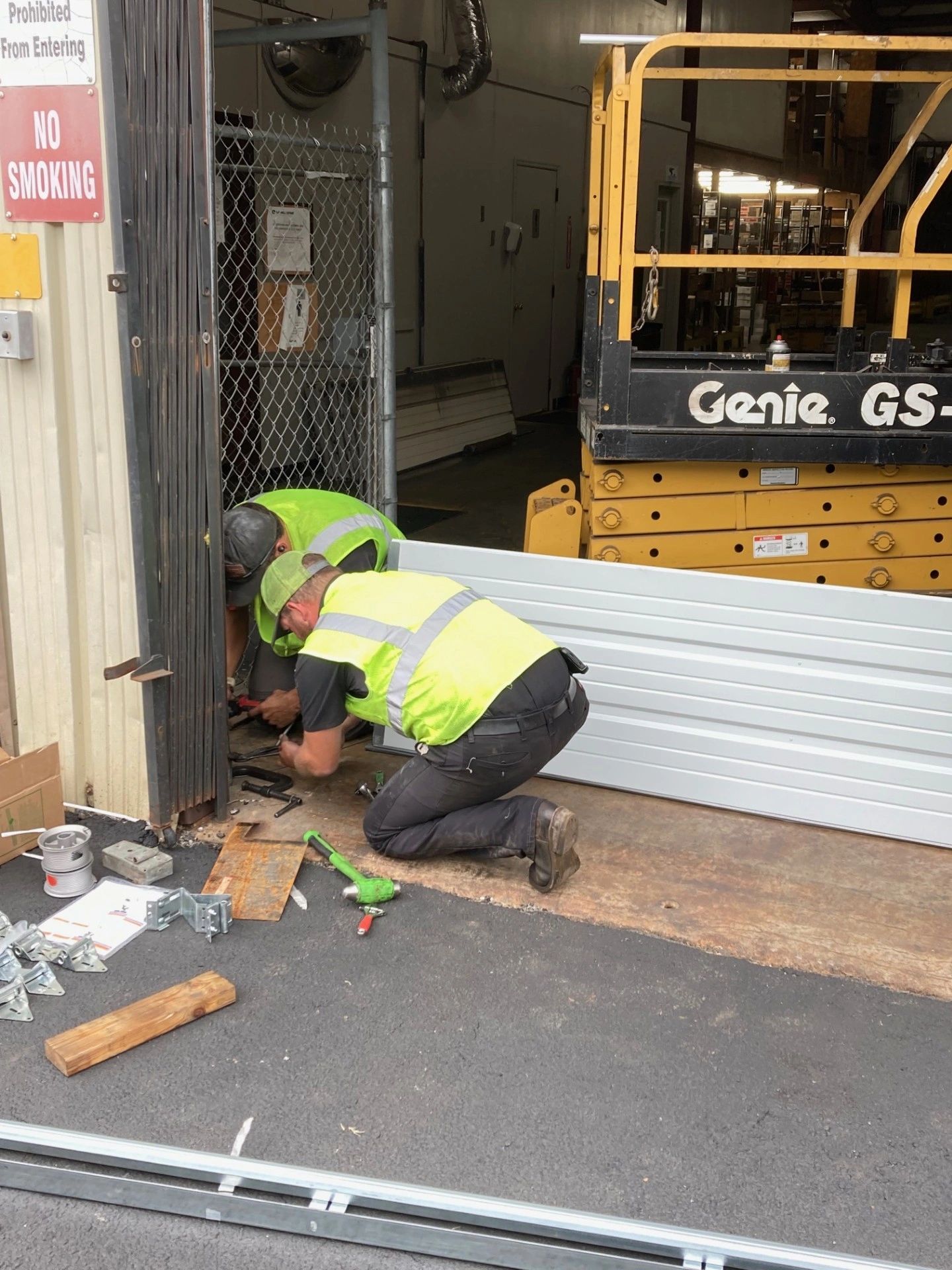 Two workers in safety vests fixing a metal panel near a warehouse entrance.