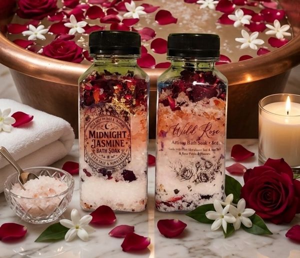 Two artisan bath soak bottles surrounded by rose petals and a lit candle.