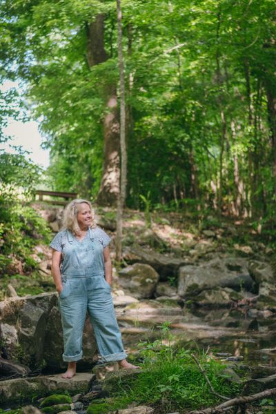 A woman in overalls standing barefoot on rocks in a lush forest.