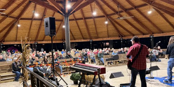 Musicians performing on stage in front of a seated audience inside a wooden pavilion.