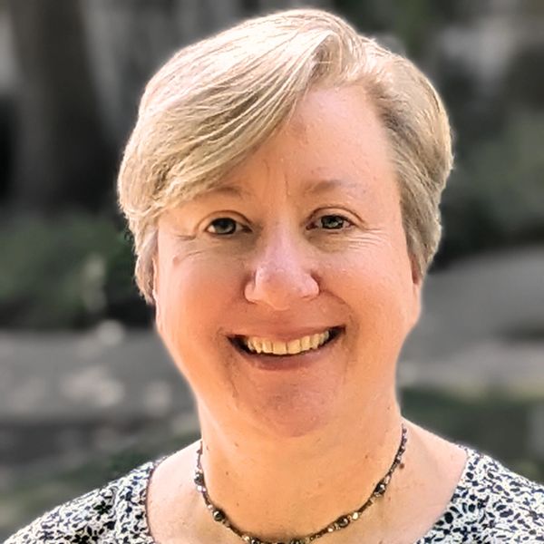 Smiling woman with short blonde hair wearing a floral blouse and a pendant necklace.