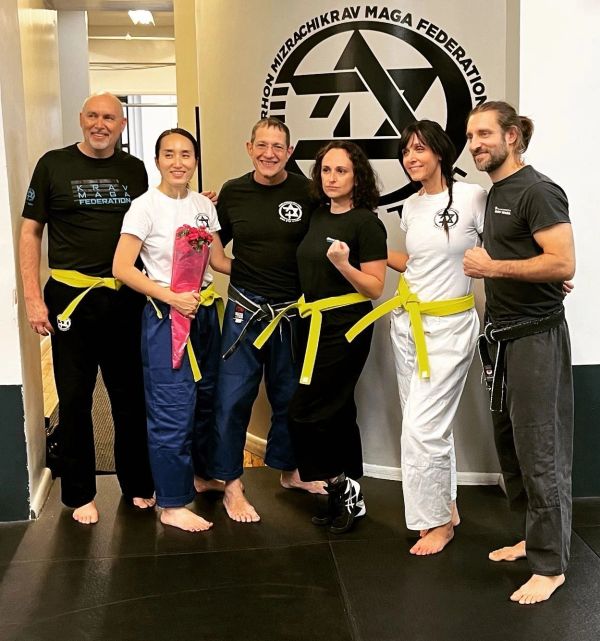Six Krav Maga practitioners posing with yellow belts and a bouquet in a martial arts gym.