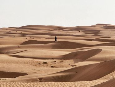 A person stands alone amid vast desert sand dunes under a clear sky.