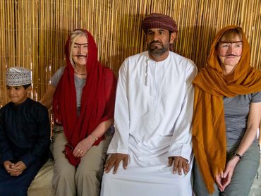 Group of people in traditional attire sitting against a bamboo wall.