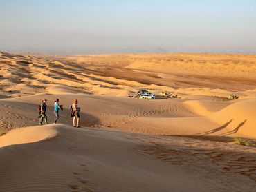 Three hikers standing on desert dunes near a campsite with vehicles and tents.