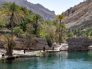 People relax by a clear water oasis amid rocky mountains and palm trees.