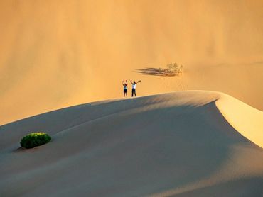 Two people celebrating atop a large sand dune in a desert landscape.