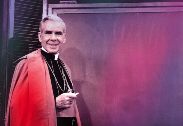 A clergyman stands smiling beside a large blank board.