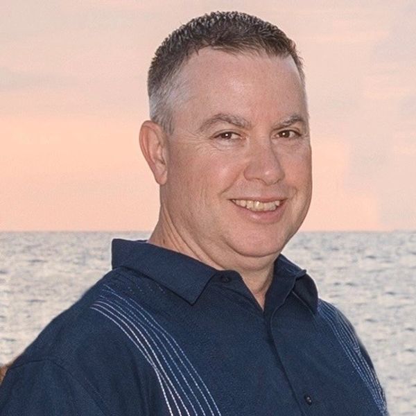 A smiling man in a blue shirt stands by the ocean at sunset.
