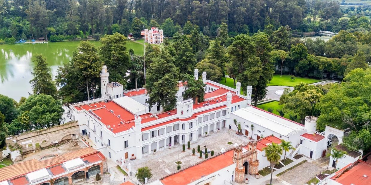 A white historic building with red roofs surrounded by trees and a lake.