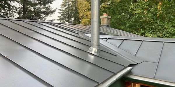 Modern metal roof with a chimney vent surrounded by trees.