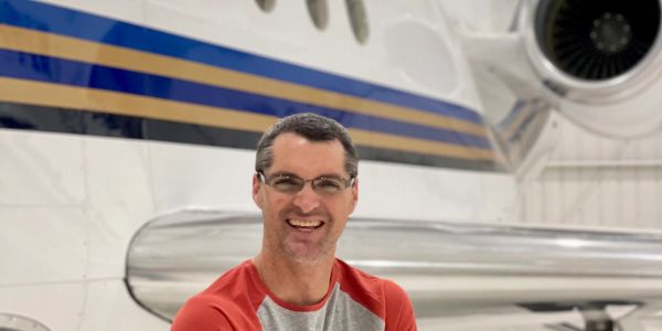 Man smiling in front of a private jet inside a hangar.