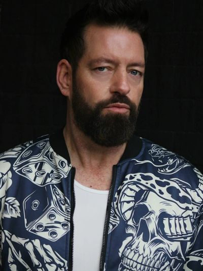 Man with a beard wearing a patterned jacket and white shirt against a dark background.