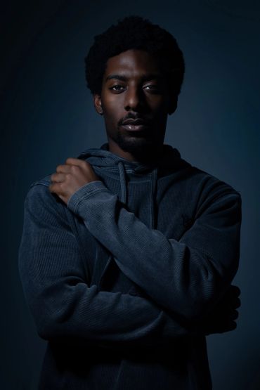 Man in dark hoodie with crossed arms in low light.