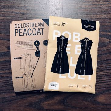 Sewing patterns for a Goldstream peacoat and a Robe Bleu dress on a wooden table.